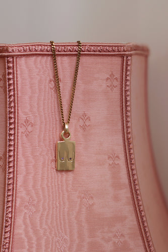 A gold-plated pendant necklace with a chain, attached to a pink textured surface.