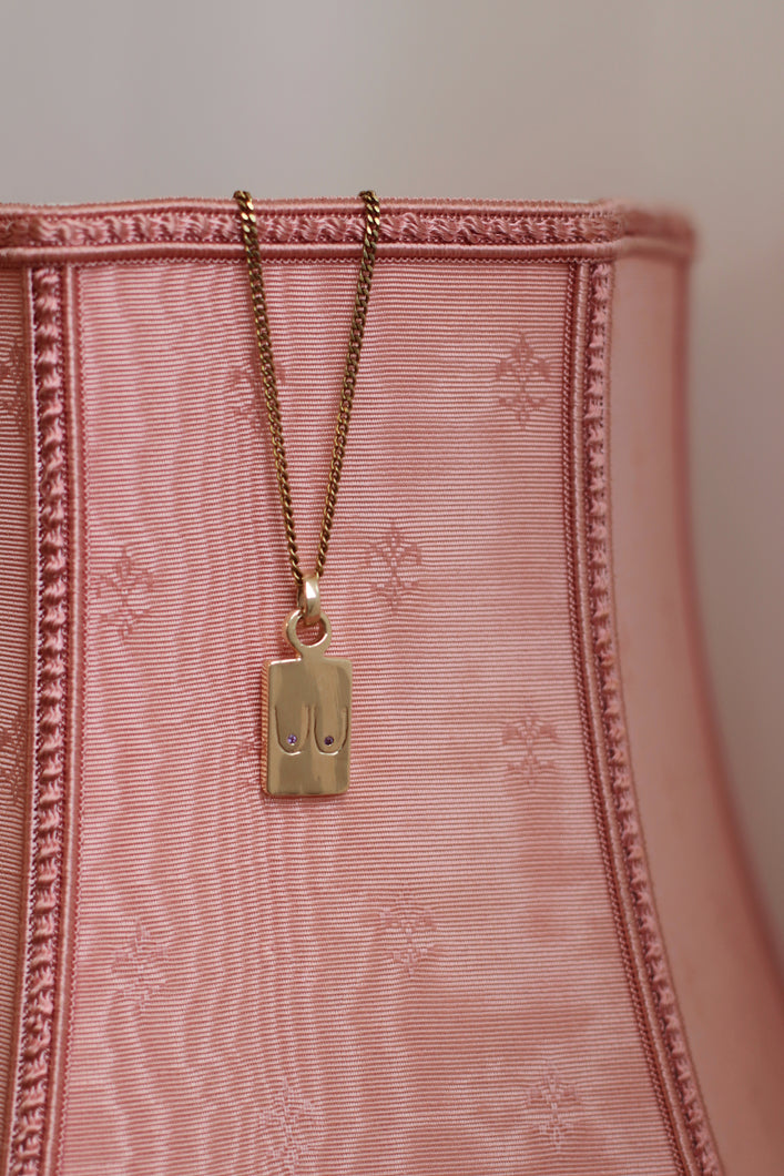 A gold-plated dainty pendant necklace with a chain, attached to a pink textured surface.