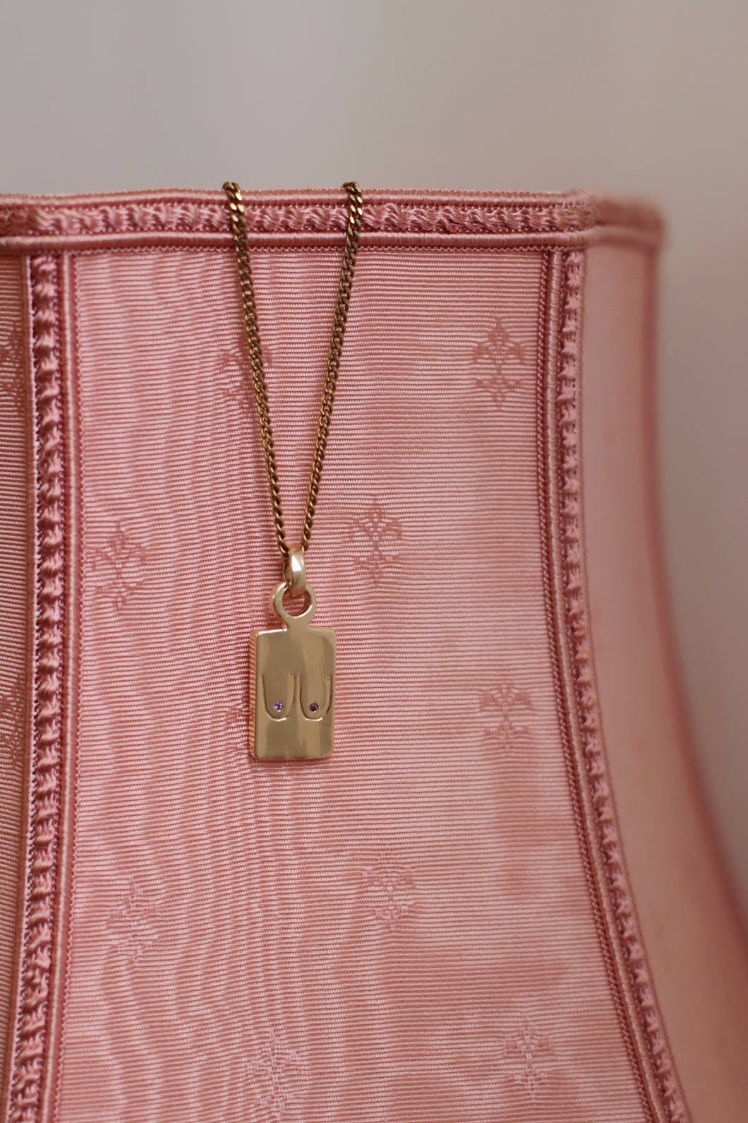 A gold-plated pendant necklace with a chain, attached to a pink textured surface.