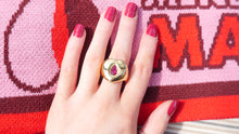 Load image into Gallery viewer, Hand wearing a gold ring with a red gemstone against a pink and red patterned background that says Menstruation Matters Belfast Scarf

